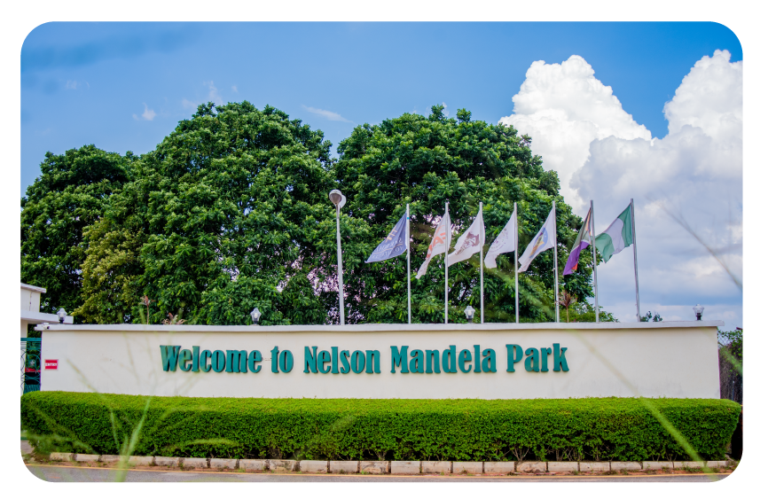 A bright sky with lush green trees by a cream coloured fence and Nelson Mandela Park boldly written on it